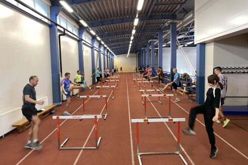 Grupa cilvēku vieglatlētikas manēžā veic stiepšanās vingrinājumus ar zemām "Nordic Sport" barjerām. Sportisti paceļ kājas pāri barjerām uz sarkanā skrejceļa, kas izvietotas divās rindās.