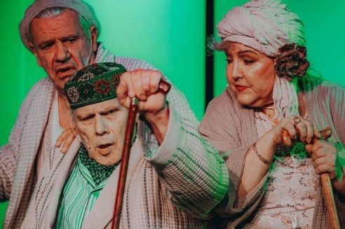 Trīs seniori – divi vīrieši un viena sieviete – tērpti halātos un ar spieķiem, sēž uz zaļi apgaismotas skatuves. Vīrietis priekšplānā, zaļā rakstainā cepurē, runā vai dzied ar atvērtu muti. Aiz viņa kreisajā pusē vīrietis ar noraizējušos izteiksmi, bet labajā pusē sieviete turbānā ar ziedu skatās uz leju ar nopietnu sejas izteiksmi.