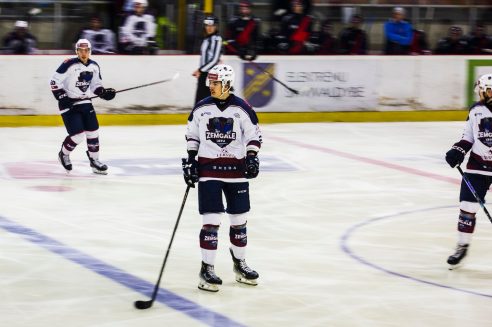 Hokejists baltā "ZEMGALE LBTU" kreklā un tumšzilos šortos stāv uz ledus ar hokeja nūju, skatoties pa labi. Uz viņa krekla redzams arī uzraksts "ŠKODA". Fonā redzami citi hokejisti, tiesnesis un aizsargapmales ar izplūdušu uzrakstu, kas daļēji salasāms kā "ELEKTREMU SWAŁDYBĘ".