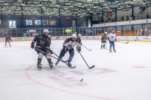 Divi hokejisti pilnā ekipējumā, viens tumšā un otrs gaišā formā, slido ar nūjām uz ledus hallē, pie ripas. Fonā redzami citi spēlētāji un skatītāji tribīnēs. Uz tablo redzams 16:10. Pie laukuma malām ir reklāmas ar tekstiem "OPTI BET", "agro24.lv", "AR AGRO", "TEAMSport.lv", "START" un "CCM".