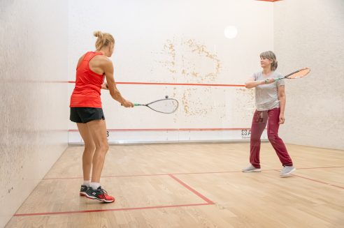 Divas sievietes spēlē skvošu kortā. Sieviete sarkanā topā sit bumbiņu ar raketi, bet sieviete pelēkā topā ir gatava spēlei. Kortā ir baltas sienas ar sarkanu līniju un koka grīda.