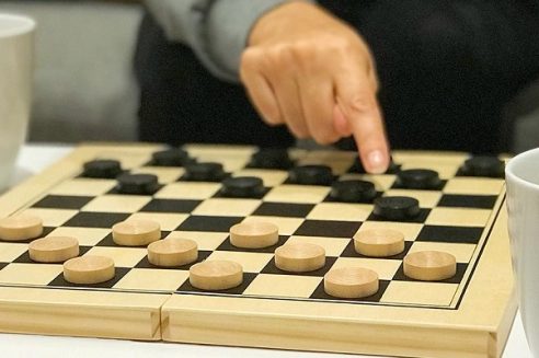 Koka dambretes spēle ar gaišiem un melniem kauliņiem. Roka pārvieto melnu kauliņu pa dēlīti. Malās redzamas baltas krūzes.