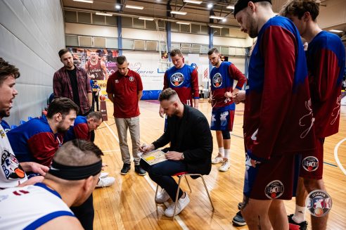Basketbola treneris sēž uz krēsla un zīmē uz taktikas dēļa, kamēr apkārt stāv vai tup spēlētāji ziliredzējos "JELGAVA BZK" formastērpos ar briežu ragu attēliem, uzmanīgi klausoties. Fons: sporta zāle ar basketbola laukumu un sienas plakātiem, tostarp ar uzrakstu "LATVIJA 3".